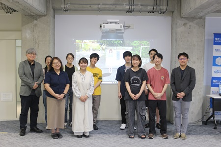 前期最終回ゼミ：完成した作品をバックに全員での集合写真 後列左端：小宮路雅博データサイエンス教育研究センター長 前列左端：小林真由美文芸学部教授 後列右端：此木啓人氏（JP UNIVERSE社ディレクター）